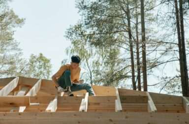 A construction worker skillfully building a wooden roof framework surrounded by tall trees.