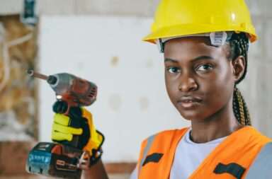 Portrait of a handywoman wearing a hardhat and reflective vest, holding a drill in a construction setting.